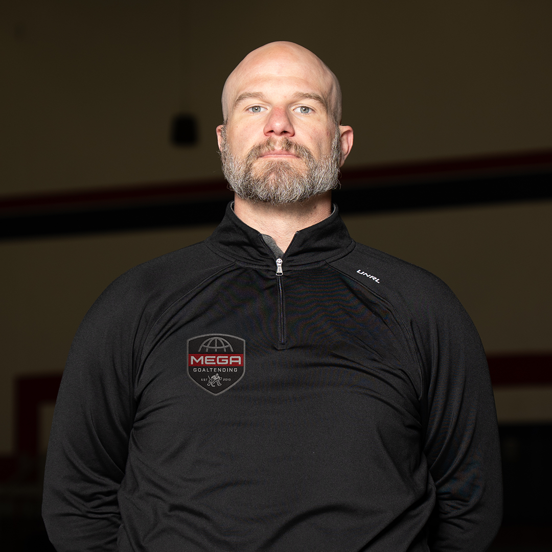 Bald, bearded man stands facing camera, arms behind back, wearing a black zip-up pullover in an indoor gym or rink.