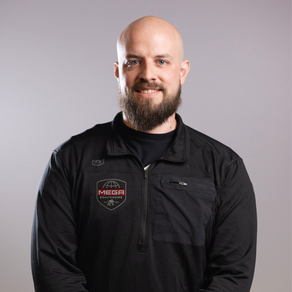 Bald, bearded man stands facing camera, arms behind back, wearing a black zip-up pullover in an indoor gym or r