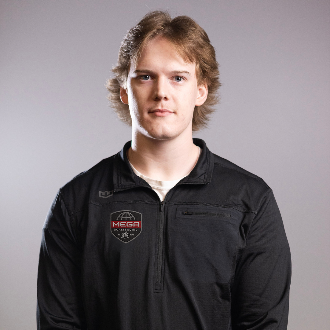 Young man looking directly at camera, wearing black zip-up jacket with patch reading "MEGA GOALTENDING", neutral gray studio background.