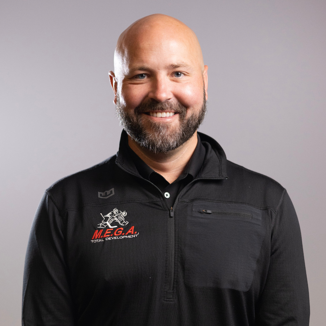 Bearded bald man smiles at camera, wearing black zip jacket against neutral gray background; logo reads: "M.E.G.A. TOTAL DEVELOPMENT".