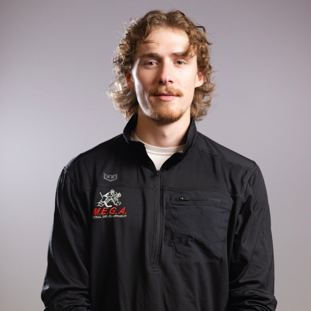 Young man faces camera with neutral expression, wearing black jacket embroidered "M.E.G.A. TOTAL DEVELOPMENT" against plain gray studio backdrop.