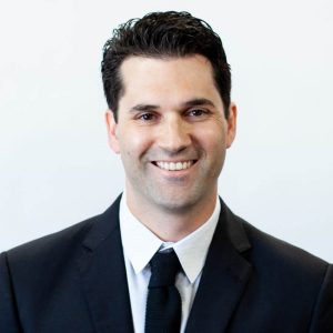 Man smiling at camera, wearing a dark suit and tie, posed against a plain light-colored background.